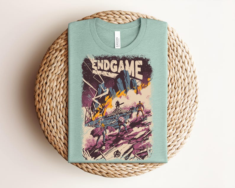 Pu&ograve; includere: Una t-shirt girocollo verde chiaro con una stampa grafica che riporta il testo ENDGAME in alto. L'illustrazione raffigura una scena di battaglia con diverse figure, fulmini, fuoco e detriti nei toni del viola, arancione e blu scuro.