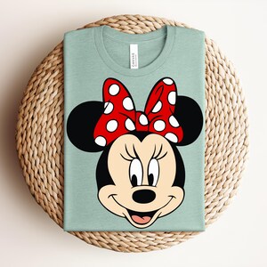 May include: A light green t-shirt featuring a large graphic of Minnie Mouse's face. The design includes her black ears, a red bow with white polka dots, and a smiling face. The shirt is folded on a woven, circular mat.