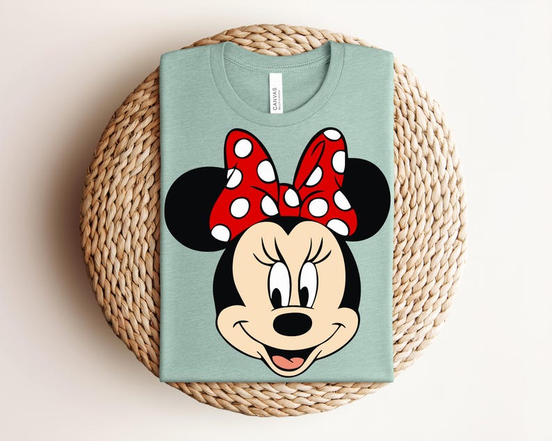 May include: A light green t-shirt featuring a large graphic of Minnie Mouse's face. The design includes her black ears, a red bow with white polka dots, and a smiling face. The shirt is folded on a woven, circular mat.