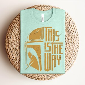 May include: A light mint green short-sleeved t-shirt featuring a distressed gold graphic of a helmet on the left and the text THIS IS THE WAY on the right. The shirt has a crew neckline and a white brand tag inside the collar area.