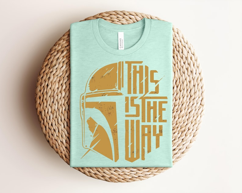 May include: A light mint green short-sleeved t-shirt featuring a distressed gold graphic of a helmet on the left and the text THIS IS THE WAY on the right. The shirt has a crew neckline and a white brand tag inside the collar area.