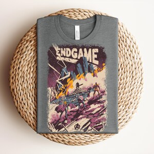 Pu&ograve; includere: Una t-shirt grigio scuro a girocollo con una stampa grafica che riporta il testo ENDGAME. L'illustrazione raffigura una scena di battaglia con figure stilizzate, fulmini, fuoco e detriti in tonalit&agrave; viola, arancione e blu, con un logo.