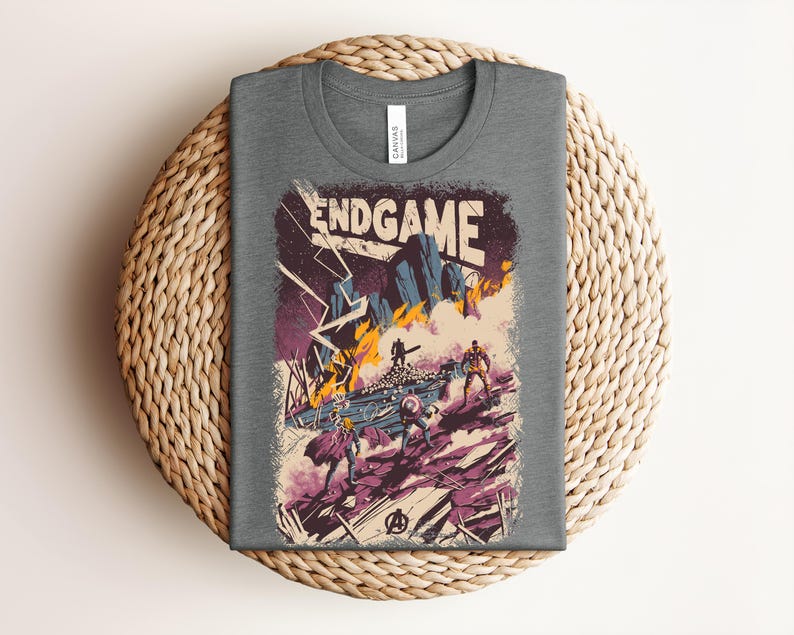 Pu&ograve; includere: Una t-shirt grigio scuro a girocollo con una stampa grafica che riporta il testo ENDGAME. L'illustrazione raffigura una scena di battaglia con figure stilizzate, fulmini, fuoco e detriti in tonalit&agrave; viola, arancione e blu, con un logo.