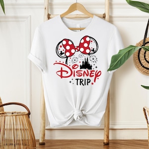 May include: White t-shirt featuring a Minnie Mouse bow with red polka dots and black mouse ears. The shirt has the words "Disney Trip" in red and black, with a castle silhouette and star accents. The shirt is hanging on a wooden hanger.