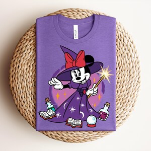 May include: A purple t-shirt featuring a cartoon Minnie Mouse dressed as a witch, holding a wand with a starburst. She wears a witch hat with a red bow and a purple dress with stars. Books, potions, and a crystal ball are also in the image.