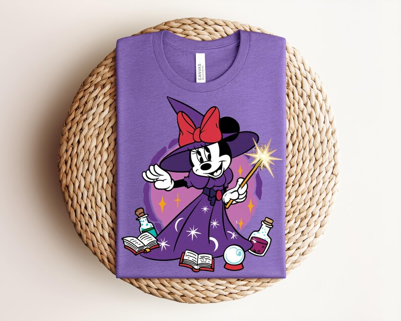 May include: A purple t-shirt featuring a cartoon Minnie Mouse dressed as a witch, holding a wand with a starburst. She wears a witch hat with a red bow and a purple dress with stars. Books, potions, and a crystal ball are also in the image.