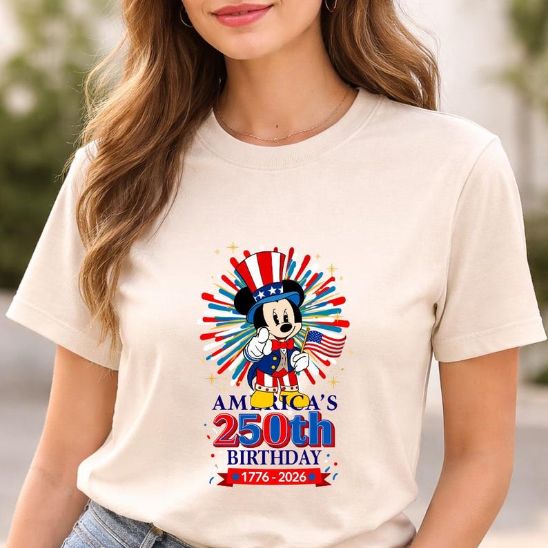 May include: Cream-colored t-shirt featuring a graphic of Mickey Mouse in a patriotic outfit, holding a flag. The design celebrates America's 250th birthday, with text reading "AMERICA'S 250th BIRTHDAY 1776-2026" in red and blue.