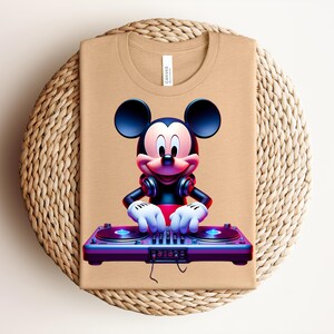 May include: Tan t-shirt featuring a graphic of Mickey Mouse as a DJ, wearing headphones and operating a turntable. The design is vibrant with red, blue, and purple hues. The shirt is folded on a woven, circular surface.