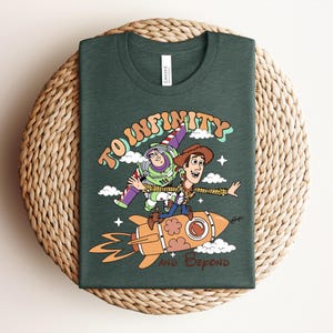 May include: A dark green t-shirt featuring a graphic print of Buzz Lightyear and Woody riding a rocket. The text reads TO INFINITY AND BEYOND in a retro font style. The design includes small white clouds and stars surrounding the characters.