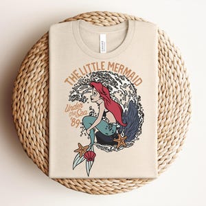 May include: A beige t-shirt featuring a graphic of a red-haired mermaid with a teal tail sitting on a rock in front of a large wave. The text reads THE LITTLE MERMAID and Under the Sea '89. Two starfish and a pink seashell are at the bottom.