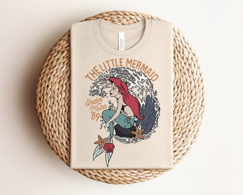 May include: A beige t-shirt featuring a graphic of a red-haired mermaid with a teal tail sitting on a rock in front of a large wave. The text reads THE LITTLE MERMAID and Under the Sea '89. Two starfish and a pink seashell are at the bottom.