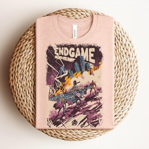 Pu&ograve; includere: Una t-shirt color pesca chiaro con una stampa grafica che riporta il testo ENDGAME. L'illustrazione raffigura una scena di battaglia con figure stilizzate, fulmini e fuoco in tonalit&agrave; viola, blu, giallo e nero, con un logo Avengers in basso.