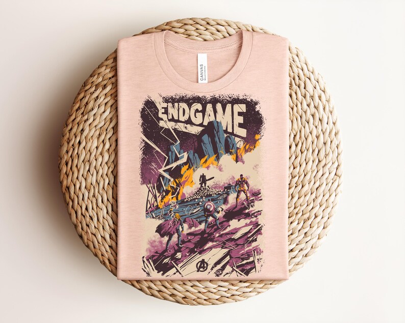 Pu&ograve; includere: Una t-shirt color pesca chiaro con una stampa grafica che riporta il testo ENDGAME. L'illustrazione raffigura una scena di battaglia con figure stilizzate, fulmini e fuoco in tonalit&agrave; viola, blu, giallo e nero, con un logo Avengers in basso.