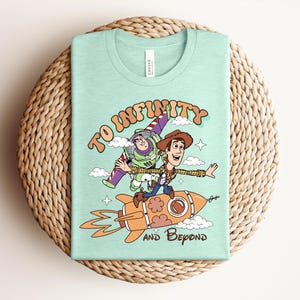 May include: A light mint green t-shirt featuring a graphic print of two cartoon characters, one in a space suit and one in a cowboy outfit, riding a rocket ship. The text "TO INFINITY AND BEYOND" is printed in a brown, stylized font around the characters.