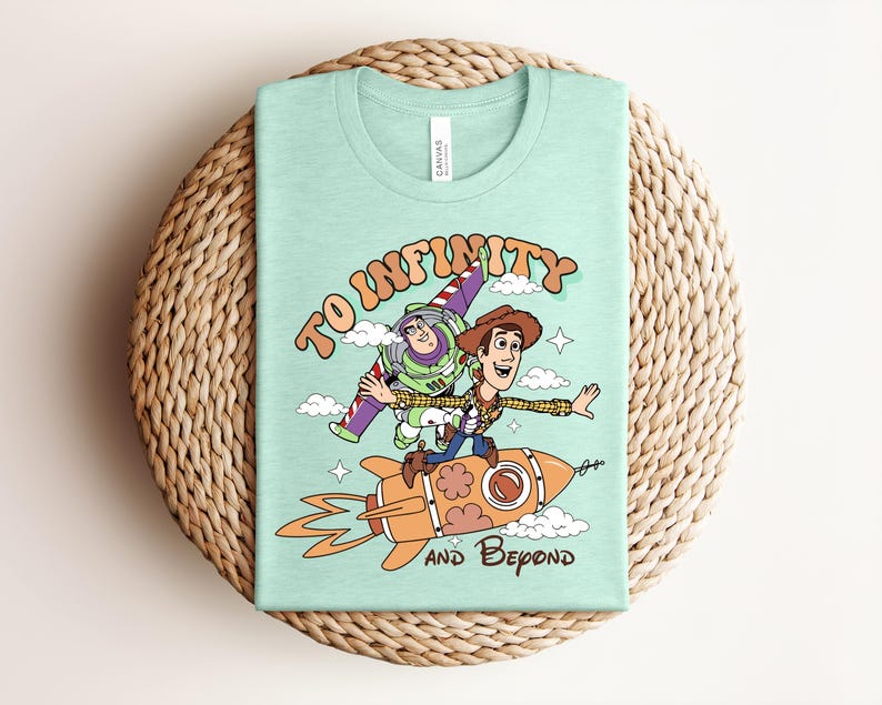 May include: A light mint green t-shirt featuring a graphic print of two cartoon characters, one in a space suit and one in a cowboy outfit, riding a rocket ship. The text "TO INFINITY AND BEYOND" is printed in a brown, stylized font around the characters.
