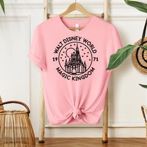 May include: A light pink t-shirt with a black graphic of the Walt Disney World logo, featuring a castle, moon, and stars. The text reads "Walt Disney World Magic Kingdom 1971". The shirt is knotted at the waist and hangs on a wooden hanger.