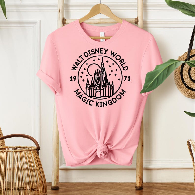 May include: A light pink t-shirt with a black graphic of the Walt Disney World logo, featuring a castle, moon, and stars. The text reads "Walt Disney World Magic Kingdom 1971". The shirt is knotted at the waist and hangs on a wooden hanger.