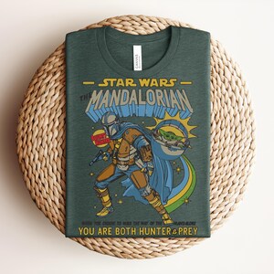 Puede incluir: Una camiseta verde oscuro con un gr&aacute;fico de The Mandalorian y Grogu. El texto dice STAR WARS THE MANDALORIAN, BOUNTY HUNTER y WHEN YOU CHOOSE TO WALK THE WAY OF THE MANDALORE YOU ARE BOTH HUNTER & PREY. El dise&ntilde;o incluye colores amarillo y azul.