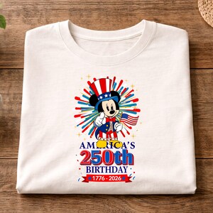 May include: White t-shirt featuring a patriotic Mickey Mouse design. Mickey is dressed in red, white, and blue, holding an American flag. The shirt celebrates America's 250th birthday, with text and fireworks graphics.