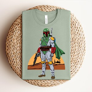 May include: Sage green t-shirt featuring a full-body illustration of a character in green, red, and gray armor, with a green cape and a blaster. The background includes a desert scene with a setting sun and a spaceship.