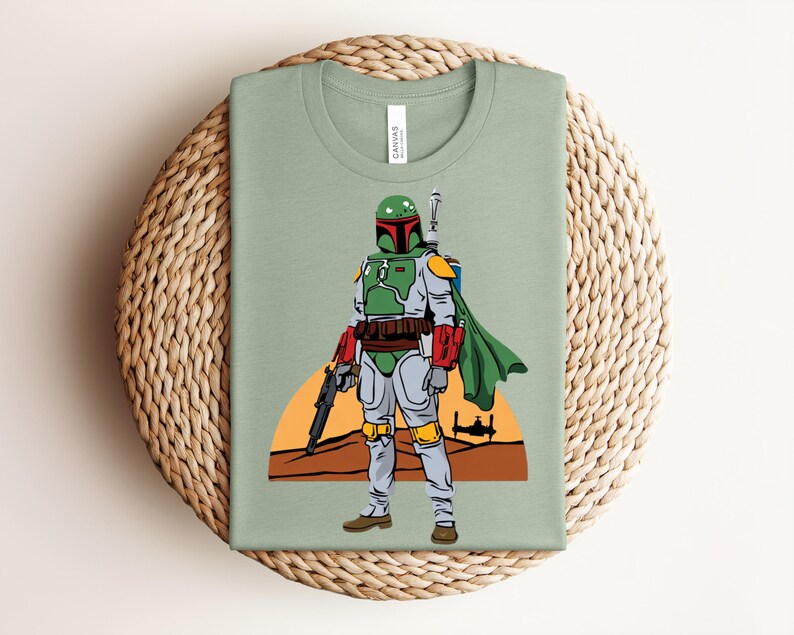 May include: Sage green t-shirt featuring a full-body illustration of a character in green, red, and gray armor, with a green cape and a blaster. The background includes a desert scene with a setting sun and a spaceship.