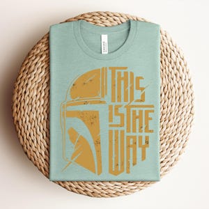 May include: A light green crew neck t-shirt featuring a distressed gold graphic of a helmet on the left side and the text "THIS IS THE WAY" in a bold, blocky font on the right side. The shirt has a white brand label inside the collar.