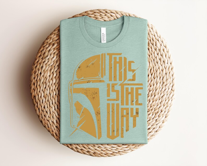 May include: A light green crew neck t-shirt featuring a distressed gold graphic of a helmet on the left side and the text "THIS IS THE WAY" in a bold, blocky font on the right side. The shirt has a white brand label inside the collar.