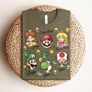May include: Olive green t-shirt featuring cartoon illustrations of Super Mario characters, including Mario, Luigi, Princess Peach, Daisy, Yoshi, and Toad. The words "Super Mario" and character names are written on the shirt.
