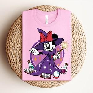 May include: Pink t-shirt featuring a cartoon Minnie Mouse dressed as a witch. Minnie wears a purple dress and hat with a red bow, holding a wand. The design includes potion bottles, a crystal ball, and open books.