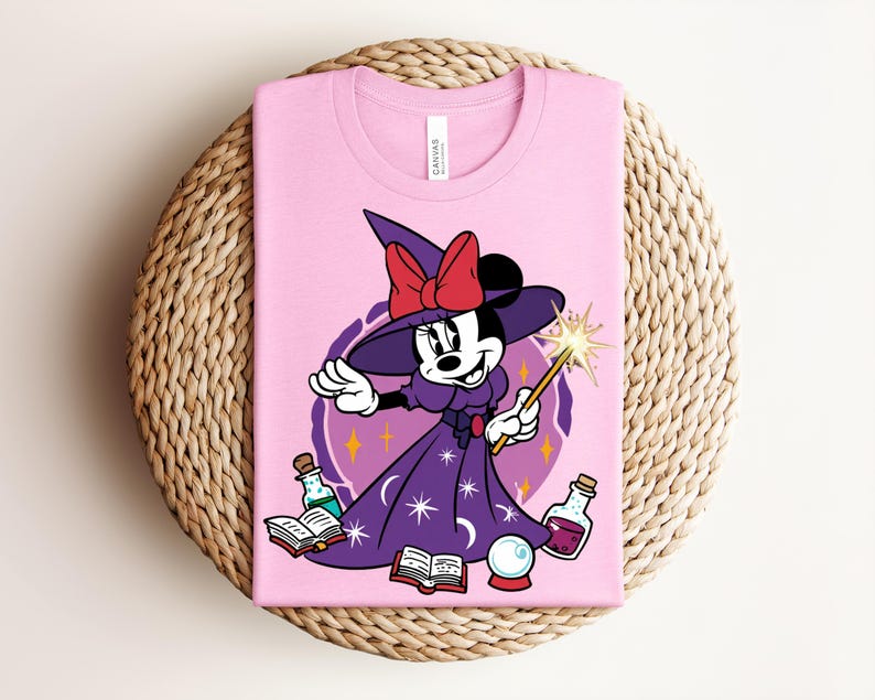 May include: Pink t-shirt featuring a cartoon Minnie Mouse dressed as a witch. Minnie wears a purple dress and hat with a red bow, holding a wand. The design includes potion bottles, a crystal ball, and open books.