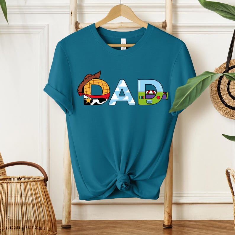 May include: Teal t-shirt with the word "DAD" in a playful design. The letter "D" features a cowboy hat, the "A" has a sky and cloud pattern, and the final "D" is designed like a toy.