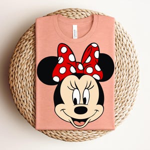May include: A peach-colored t-shirt featuring a cartoon image of Minnie Mouse. The design includes Minnie's face, black ears, and a red bow with white polka dots. The shirt is folded and placed on a woven, circular surface.