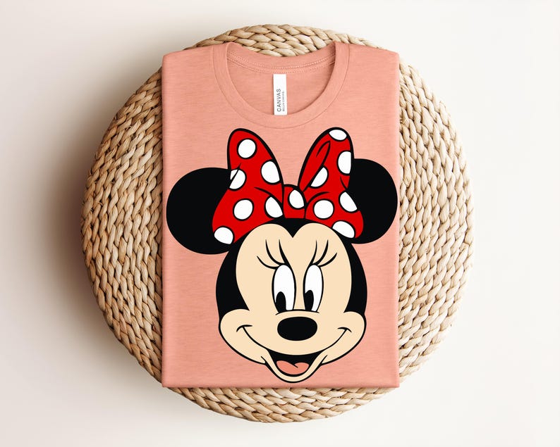 May include: A peach-colored t-shirt featuring a cartoon image of Minnie Mouse. The design includes Minnie's face, black ears, and a red bow with white polka dots. The shirt is folded and placed on a woven, circular surface.