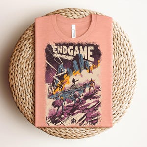 Pu&ograve; includere: Una t-shirt color pesca con un'illustrazione grafica che riporta il testo ENDGAME in alto. L'opera raffigura una scena di battaglia con diverse figure, fulmini e un terreno roccioso nei toni del viola, del blu e del giallo.