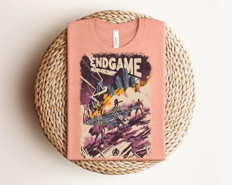 Pu&ograve; includere: Una t-shirt color pesca con un'illustrazione grafica che riporta il testo ENDGAME in alto. L'opera raffigura una scena di battaglia con diverse figure, fulmini e un terreno roccioso nei toni del viola, del blu e del giallo.