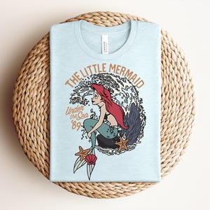 May include: A light blue t-shirt featuring a graphic of a red-haired mermaid sitting on a rock amidst ocean waves. The text reads THE LITTLE MERMAID and Under the Sea '89. Two orange starfish and a pink seashell are positioned near the mermaid's tail.
