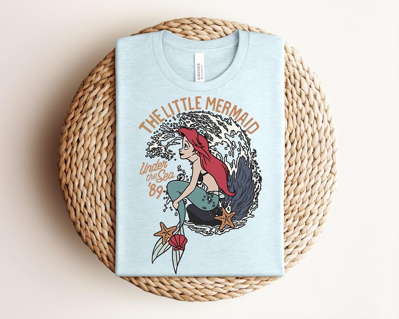 May include: A light blue t-shirt featuring a graphic of a red-haired mermaid sitting on a rock amidst ocean waves. The text reads THE LITTLE MERMAID and Under the Sea '89. Two orange starfish and a pink seashell are positioned near the mermaid's tail.