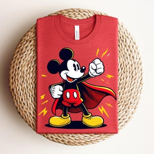 May include: Red t-shirt featuring a cartoon Mickey Mouse in a superhero pose with a red cape and yellow lightning bolts. The shirt is folded and placed on a woven, circular mat.