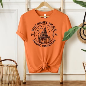 May include: Orange t-shirt with a black graphic of the Walt Disney World logo, featuring a castle, moon, and stars. The text reads "Walt Disney World Magic Kingdom 1971". The shirt is hanging on a wooden rack.