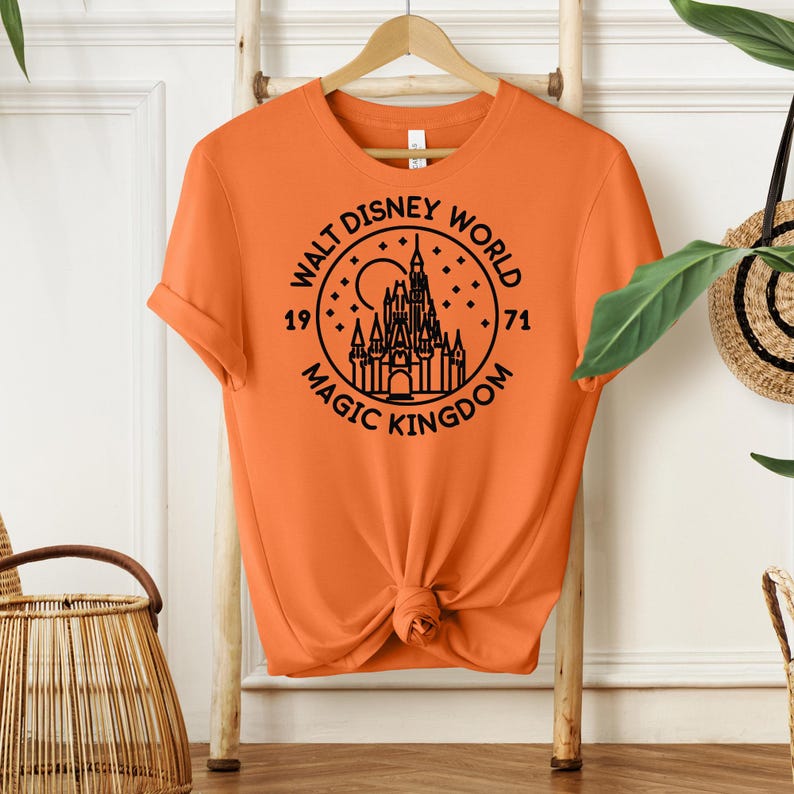 May include: Orange t-shirt with a black graphic of the Walt Disney World logo, featuring a castle, moon, and stars. The text reads "Walt Disney World Magic Kingdom 1971". The shirt is hanging on a wooden rack.