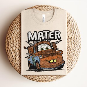 May include: A beige crew neck t-shirt featuring a graphic print of a rusty brown tow truck character with green eyes and large teeth. The word MATER is printed in large white block letters above the truck, with a black and white checkered pattern.