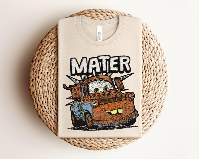 May include: A beige crew neck t-shirt featuring a graphic print of a rusty brown tow truck character with green eyes and large teeth. The word MATER is printed in large white block letters above the truck, with a black and white checkered pattern.