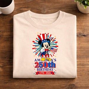 May include: Cream-colored t-shirt featuring a patriotic Mickey Mouse design. Mickey is dressed in a red, white, and blue outfit, holding an American flag. The shirt celebrates America's 250th birthday, with text reading "AMERICA'S 250th BIRTHDAY 1776-2026."