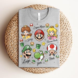 May include: A gray t-shirt featuring colorful illustrations of characters from the Super Mario video game series. The design includes Mario, Luigi, Princess Peach, Daisy, Yoshi, and Toad, along with iconic elements like coins and mushrooms.