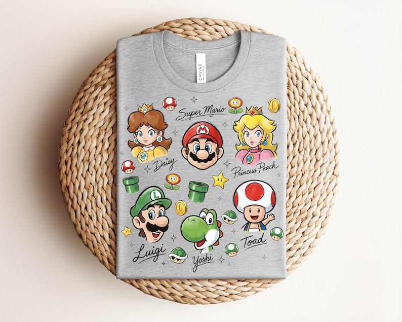 May include: A gray t-shirt featuring colorful illustrations of characters from the Super Mario video game series. The design includes Mario, Luigi, Princess Peach, Daisy, Yoshi, and Toad, along with iconic elements like coins and mushrooms.