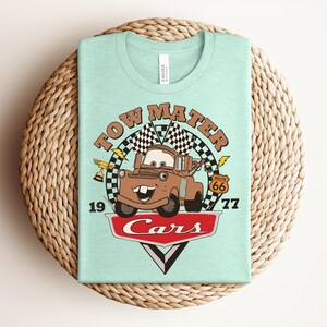 May include: A light mint green t-shirt featuring a graphic of the character Tow Mater from the movie Cars. The design includes the text "TOW MATER", "1977", "Route 66", and "Cars" with checkered flags, a trophy, and lightning bolt accents.