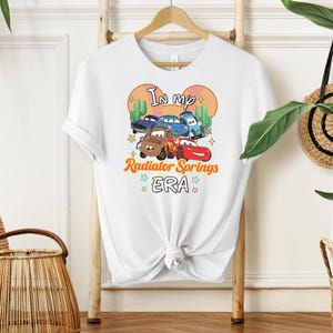 K&ouml;nnte beinhalten: Wei&szlig;es T-Shirt mit einem farbenfrohen Aufdruck von Zeichentrickautos und dem Text "In my Radiator Springs ERA". Das Design beinhaltet Kakteen und Sterne, was ein verspieltes und nostalgisches Thema schafft. Das Shirt h&auml;ngt an einem Holzgestell.
