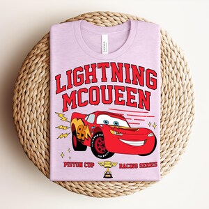 May include: A light purple t-shirt featuring a graphic of Lightning McQueen from the movie Cars. The design includes the text "LIGHTNING McQUEEN" in red, the number 95, and the words "Piston Cup Racing Series".