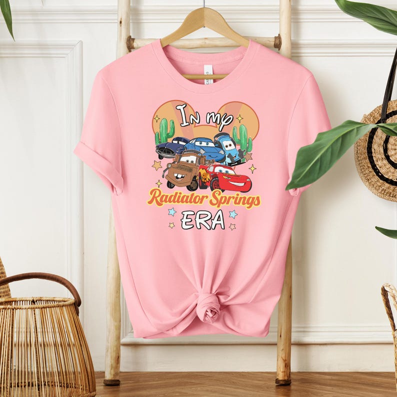 K&ouml;nnte beinhalten: Hellrosa T-Shirt mit einer Grafik von Cartoon-Autos und dem Text "In my Radiator Springs ERA". Das Design beinhaltet Kakteen und Sterne und erzeugt ein verspieltes und nostalgisches Thema.