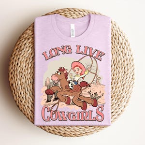 May include: A light purple t-shirt featuring a graphic of a cartoon cowgirl riding a brown horse in a desert landscape with cacti. The text "LONG LIVE COWGIRLS" is printed in a pink retro font above and below the illustration. The shirt has a crew neckline.